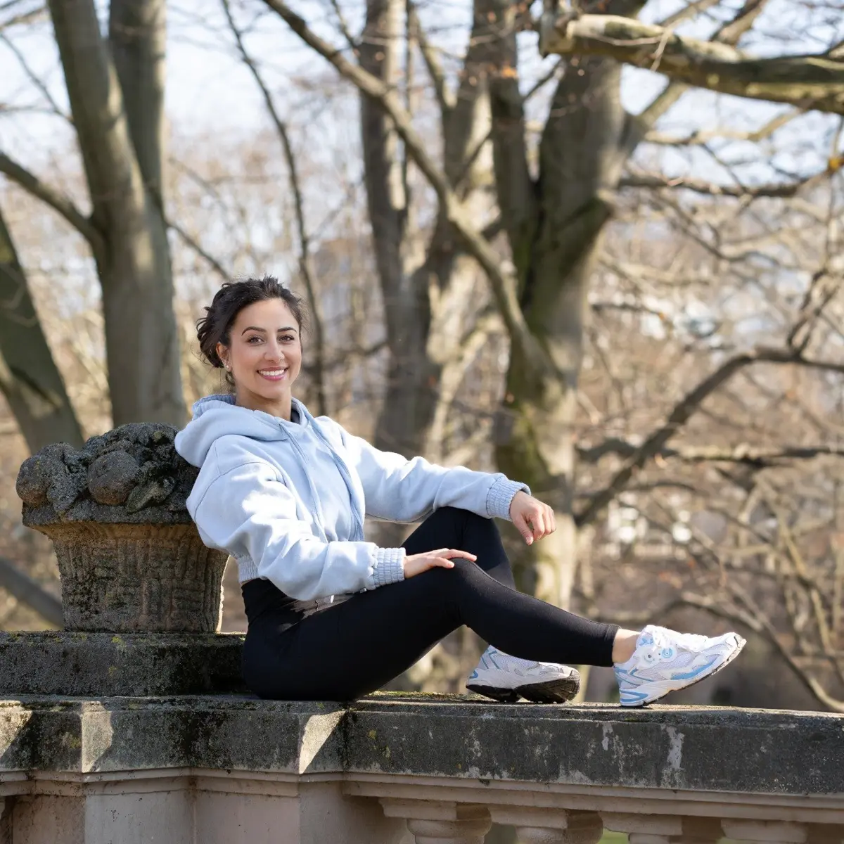 Dee in hellblauem Hoodie sitzt lächelnd auf einer Steinmauer im Park